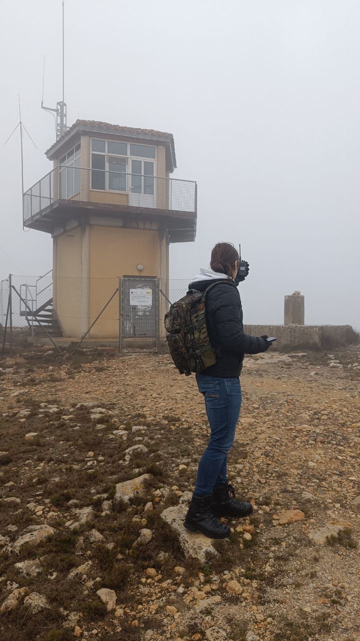 Radioaficionada con walkie y mochila junto a una torre de vigilancia entre la niebla