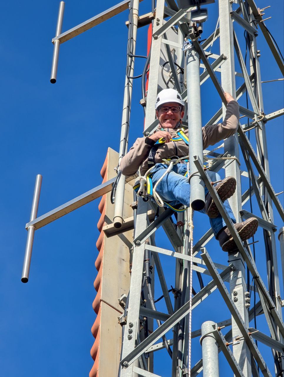 Voluntario con casco y arnés subido a una torre de antenas, con una antena yagi al fondo